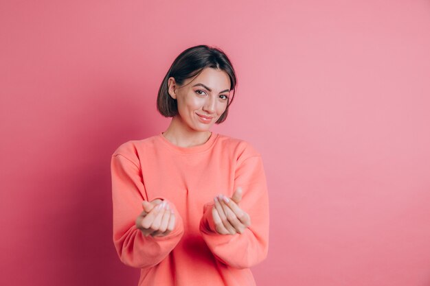 Lächelnde junge brünette Frau 20s, die lässig stehende reibende Finger zeigt, zeigt Geldgeste, die um Geld rosa Hintergrund bittet