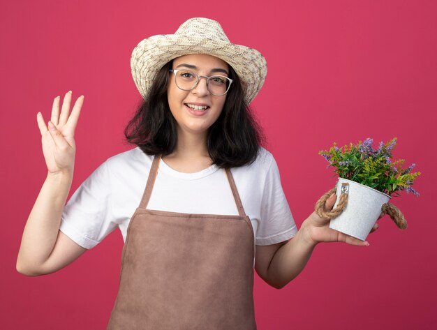 Lächelnde junge brünette Gärtnerin in optischen Gläsern und in Uniform mit Gartenhut hält Blumentopf und gestikuliert vier mit Fingern isoliert auf rosa Wand mit Kopierraum