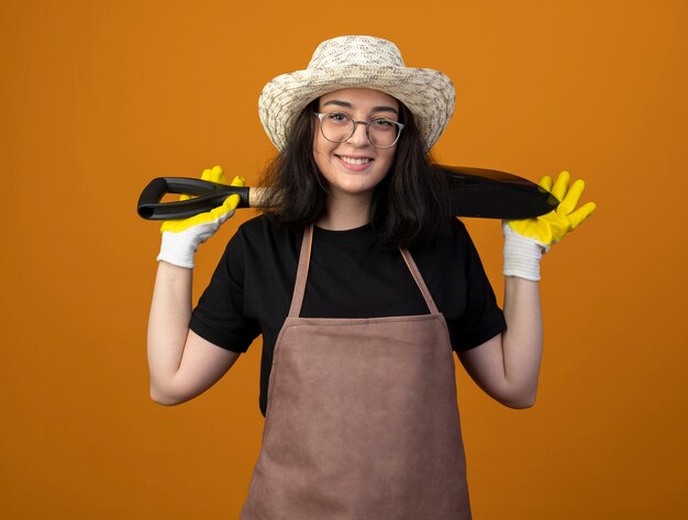 Lächelnde junge brünette Gärtnerin in optischen Gläsern und in Uniform mit Gartenhut und Handschuhen hält Spaten hinter dem Hals isoliert auf orange Wand mit Kopienraum