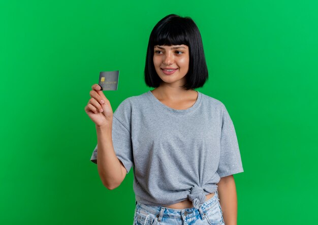 Lächelnde junge brünette kaukasische Frau hält Kreditkarte, die Seite lokalisiert auf grünem Hintergrund mit Kopienraum betrachtet