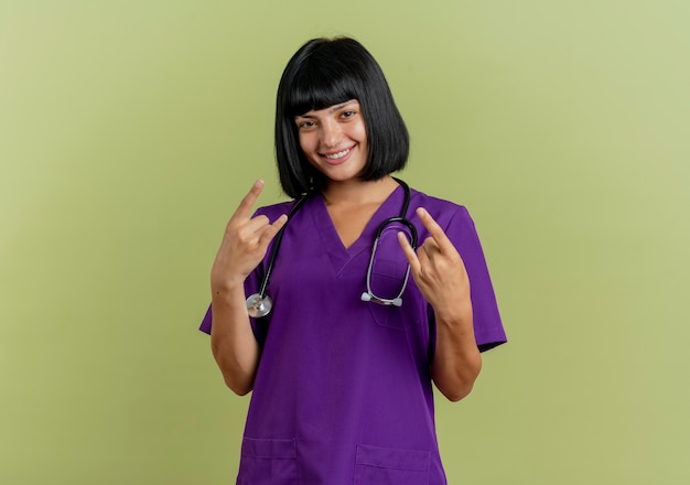 Lächelnde junge brünette Ärztin in Uniform mit Stethoskop gestikuliert Hörner Handzeichen mit zwei Händen