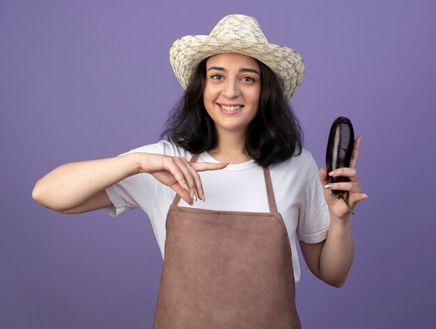 Lächelnde junge brünette weibliche Gärtnerin in Uniform, die Gartenhut trägt, hält und zeigt auf Aubergine lokalisiert auf lila Wand mit Kopienraum