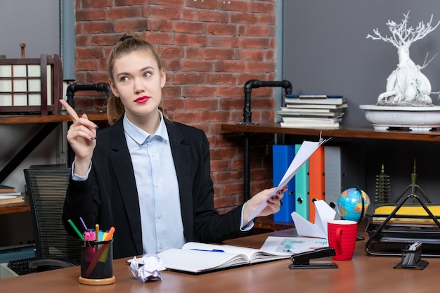 Lächelnde junge Dame, die an einem Tisch sitzt und das Dokument im Büro nach oben zeigt