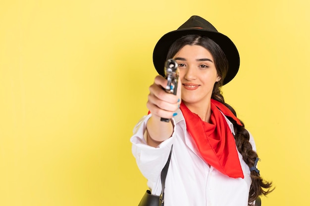 Lächelnde junge Dame, die auf gelbem Hintergrund steht und ihre Waffe auf die Kamera richtet Foto in hoher Qualität