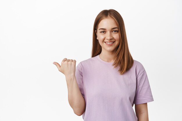 Lächelnde junge Frau, Assistentin zeigt mit dem Daumen nach links und sieht freundlich aus, zeigt die Richtung, Logo-Banner auf leerem Raum beiseite, weiße Wand