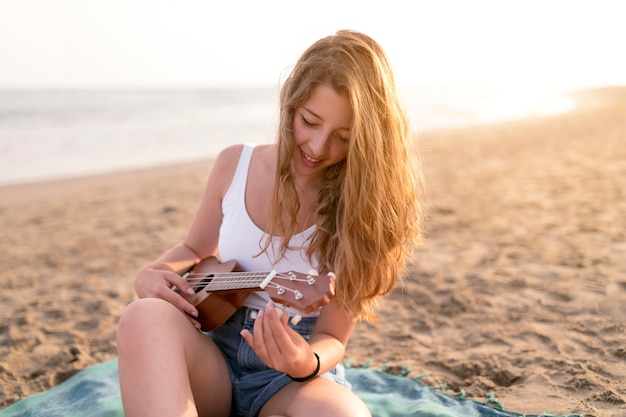 Lächelnde junge Frau, die am Strand spielt Ukulele sitzt