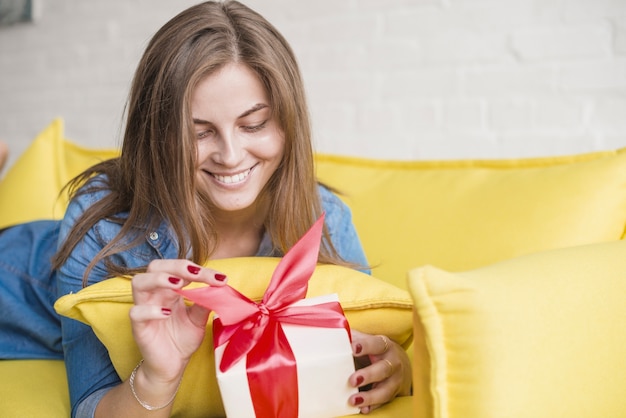 Lächelnde junge Frau, die an vom Sofa auspackt ihr Geburtstagsgeschenk liegt