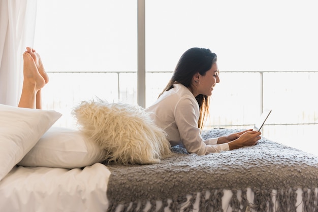 Lächelnde junge Frau, die auf Bett unter Verwendung der digitalen Tablette im Schlafzimmer liegt