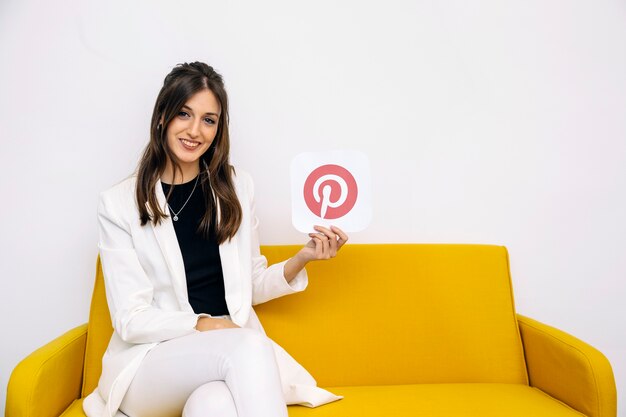 Lächelnde junge Frau, die auf dem gelben Sofa zeigt pinterest Ikone sitzt