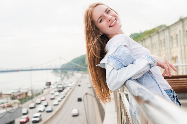 Lächelnde junge Frau, die auf dem metallischen Geländer betrachtet Kamera sich lehnt