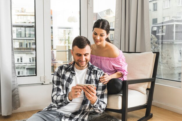 Lächelnde junge Frau, die auf dem Stuhl betrachtet ihren Freund verwendet Handy sitzt