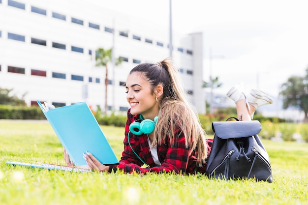 Lächelnde junge Frau, die auf den Rasen liest das Buch am Universitätsgelände legt