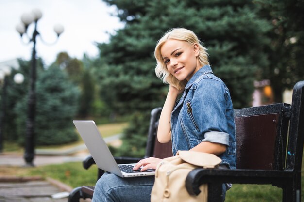 Lächelnde junge Frau, die auf der Bank sitzt und Telefon und Laptop im Herbstmorgen der Stadt benutzt