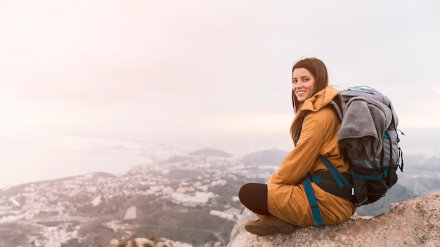 Lächelnde junge Frau, die auf die Oberseite des Berges mit ihrem Rucksack sitzt