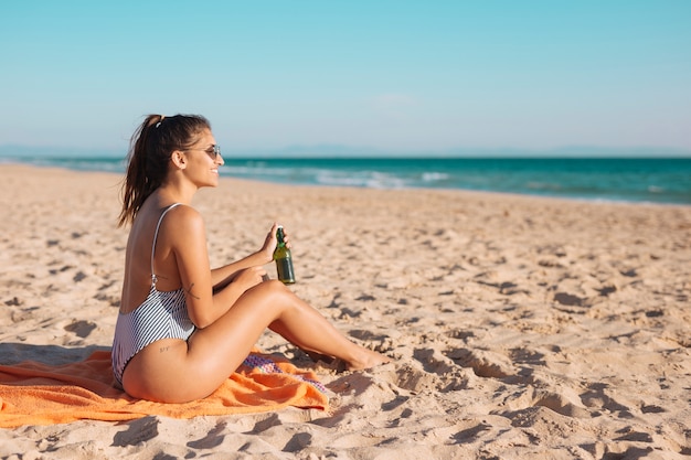 Lächelnde junge Frau, die auf Strand mit Bier sich entspannt