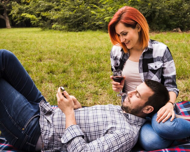 Lächelnde junge Frau, die das Weinglas betrachtet den Mann verwendet Handy im Park hält