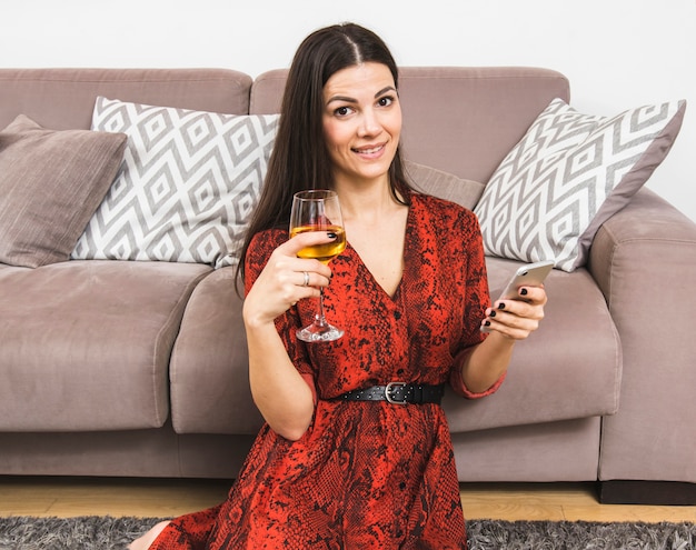 Lächelnde junge Frau, die den Handy und Weinglas in der Hand sitzen nahe dem Sofa hält