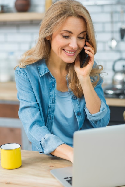 Lächelnde junge Frau, die den Laptop spricht am intelligenten Telefon verwendet