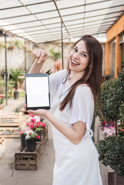 Lächelnde junge Frau, die digitale Tablette mit leerem weißem Schirm im Gewächshaus zeigt