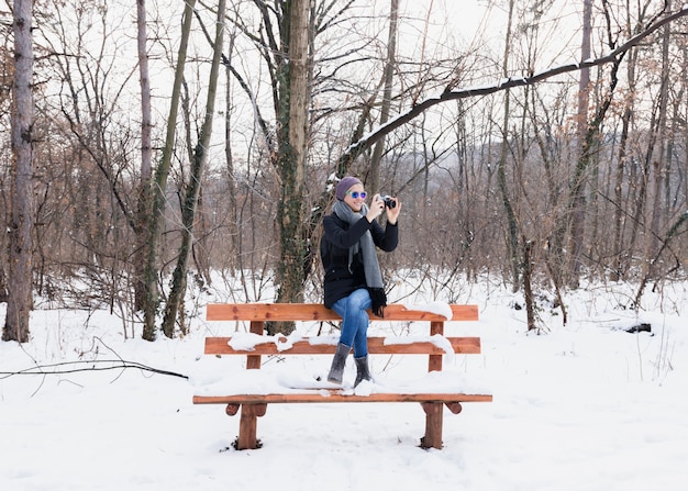 Lächelnde junge Frau, die Fotos im Winter sitzt auf Bank im Schnee macht