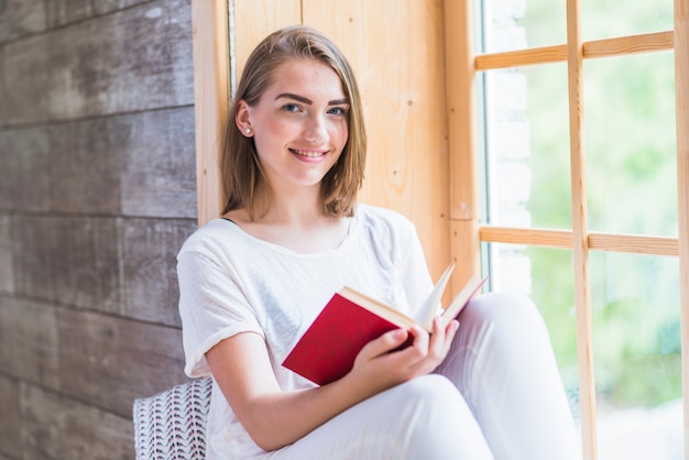 Kostenloses Foto lächelnde junge frau, die nahe fensterlesebuch sitzt