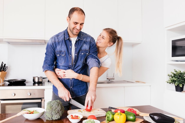 Lächelnde junge Frau, die seinen Ehemann von hinten umfasst, das Gemüse mit Messer schneidend