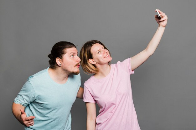 Lächelnde junge Frau, die selfie mit ihrem Freund gegen grauen Hintergrund nimmt