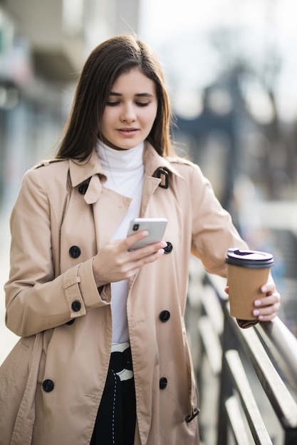 Lächelnde junge Frau im hellbraunen Mantel las Nachrichten am Telefon draußen