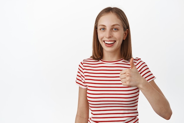 Lächelnde junge Frau, Studentin zeigt Daumen hoch, sieht zufrieden aus, empfiehlt Produkt, gefällt und stimmt zu, positives Feedback, steht gegen weiße Wand.