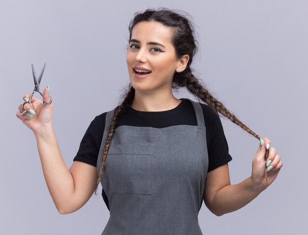 Lächelnde junge Friseurin in Uniform mit Schere und gepacktem Haar isoliert auf weißer Wand