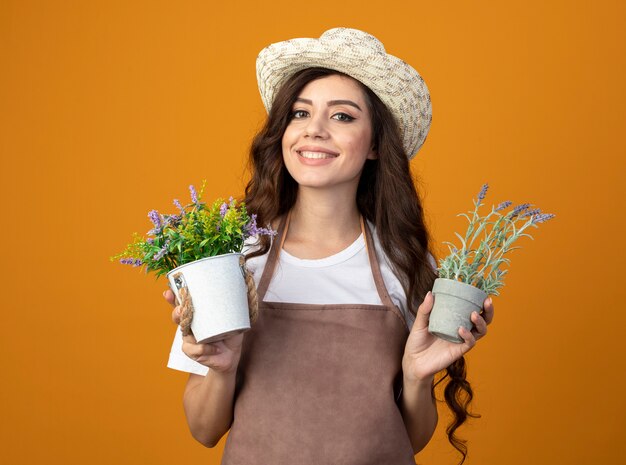 Lächelnde junge Gärtnerin in Uniform, die Gartenhut trägt, hält Blumentöpfe lokalisiert auf orange Wand mit Kopienraum