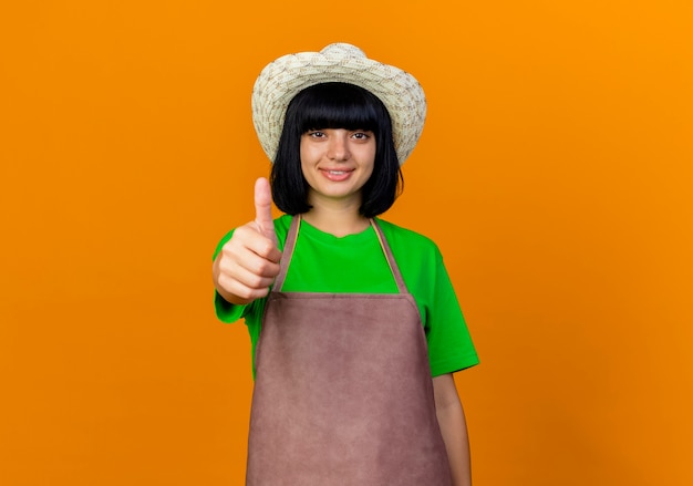 Lächelnde junge gärtnerin in uniform mit gartenhut daumen hoch