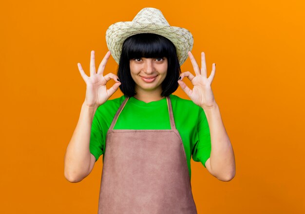 Lächelnde junge Gärtnerin in Uniform mit Gartenhut gestikuliert ok Handzeichen mit zwei Händen