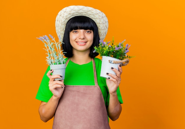 Lächelnde junge Gärtnerin in Uniform mit Gartenhut hält Blumentöpfe