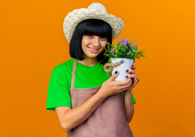 Lächelnde junge Gärtnerin in Uniform mit Gartenhut hält Blumentopf