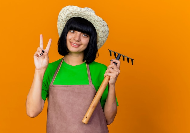 Lächelnde junge Gärtnerin in Uniform mit Gartenhut hält Rechen und Geste Sieg