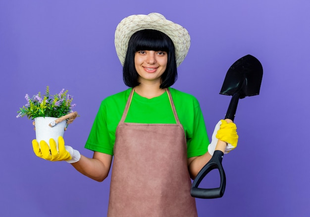 Lächelnde junge Gärtnerin in Uniform mit Gartenhut und Handschuhen hält Spaten
