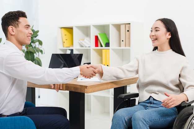 Lächelnde junge Geschäftsfrau, die auf dem Rollstuhl rüttelt Hände mit Geschäftsmann im Büro sitzt