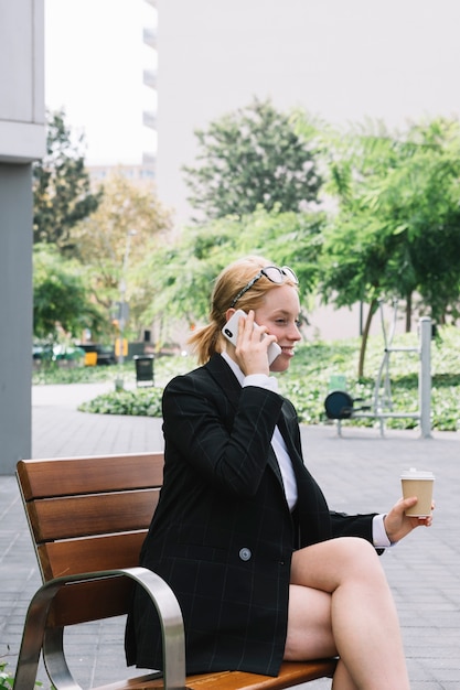 Lächelnde junge Geschäftsfrau, die auf der Bank hält Mitnehmerkaffeetasse spricht am Handy sitzt