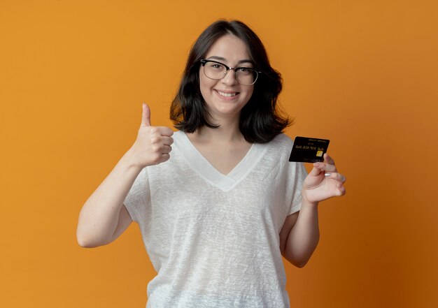 lächelnde junge hübsche Frau, die Brille trägt, Daumen zeigt und Kreditkarte hält