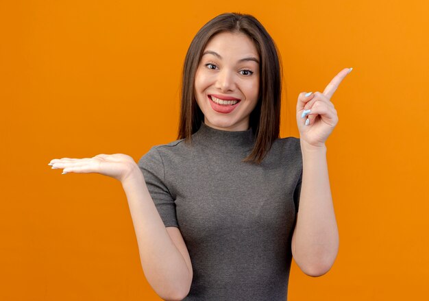 Lächelnde junge hübsche Frau, die leere Hand zeigt und auf Seite lokalisiert auf orange Hintergrund zeigt
