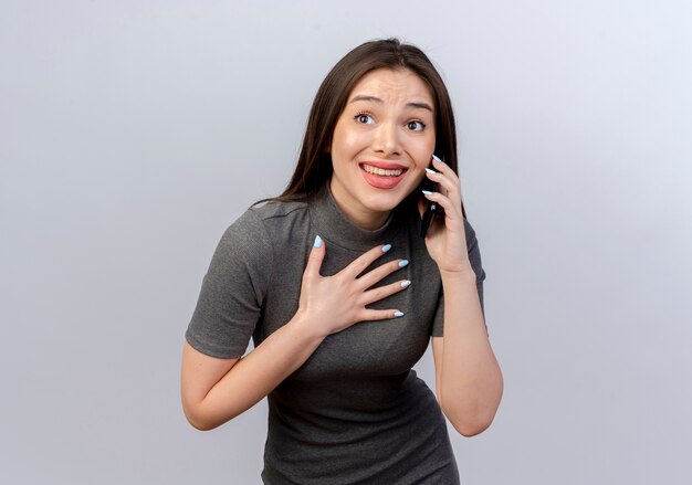 Lächelnde junge hübsche Frau, die Seite betrachtet, die Hand auf Brust setzt, die am Telefon spricht, lokalisiert auf weißem Hintergrund mit Kopienraum