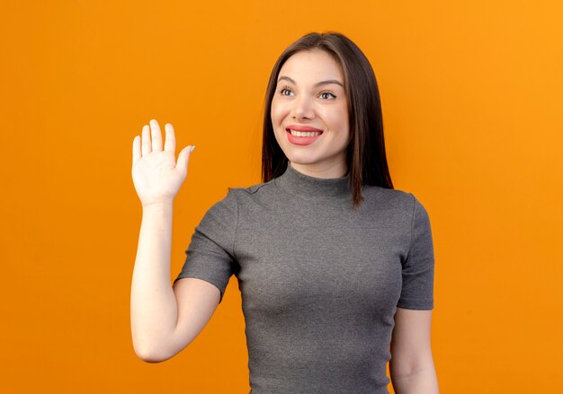 Lächelnde junge hübsche Frau, die Seite betrachtet und Hallo lokalisiert auf orange Hintergrund gestikuliert