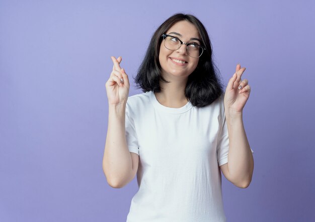 lächelnde junge hübsche Frau mit Brille, die nach oben schaut und gekreuzte Finger gestikuliert