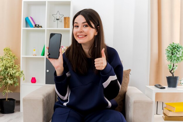 Lächelnde junge hübsche kaukasische Frau, die auf Sessel in entworfenem Wohnzimmer sitzt, zeigt Handy und zeigt Daumen hoch schauend