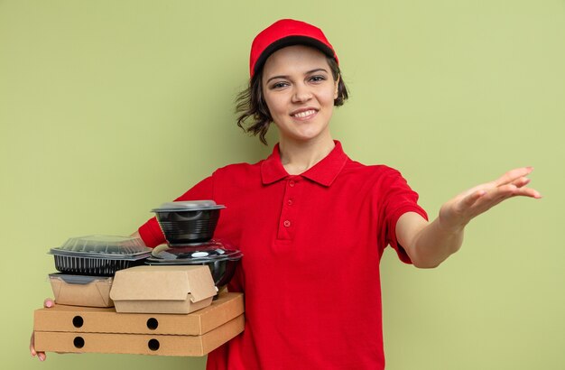 Lächelnde junge hübsche Lieferfrau, die Lebensmittelbehälter mit Verpackung auf Pizzakartons hält und ihre Hand offen hält