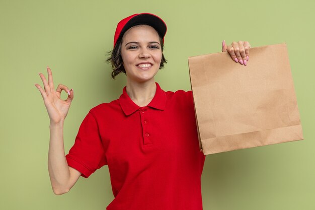 Lächelnde junge hübsche Lieferfrau, die Papierverpackungen für Lebensmittel hält und ein OK-Zeichen gestikuliert