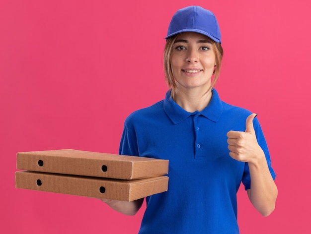 Lächelnde junge hübsche Lieferfrau in Uniform Daumen hoch und hält Pizzaschachteln isoliert auf rosa Wand
