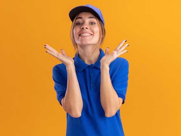 Lächelnde junge hübsche Lieferfrau in Uniform hält Hände offen auf orange Wand offen