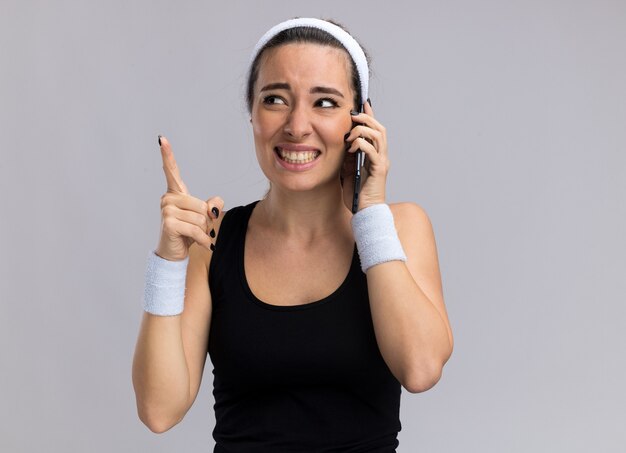 Lächelnde junge hübsche sportliche Frau mit Stirnband und Armbändern, die am Telefon spricht und auf die Seite schaut, die nach oben zeigt, isoliert auf weißer Wand mit Kopierraum